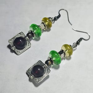 May include: A pair of handcrafted earrings featuring dark red, round beads set in ornate silver-toned square frames. The earrings also include green and yellow glass beads, and silver-toned ear wires.