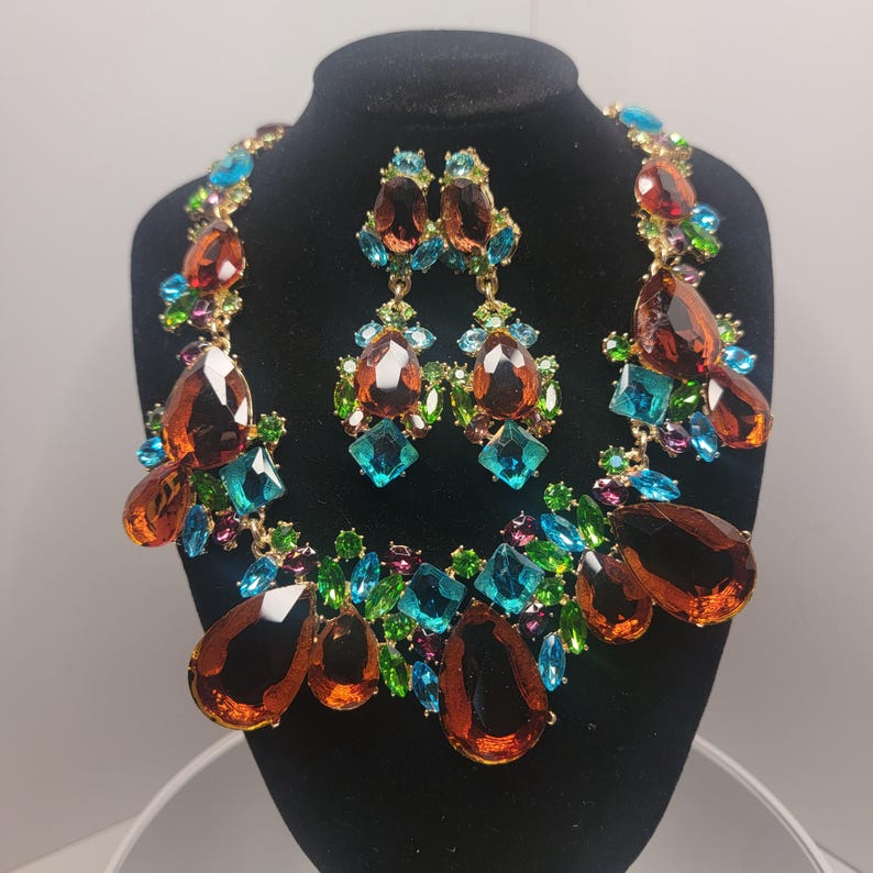 May include: A jewelry set consisting of a statement necklace and matching drop earrings. The pieces feature large amber-colored teardrop crystals accented by smaller bright blue, green, and purple faceted stones set in a gold-tone metal frame.