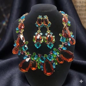 May include: A jewelry set consisting of a necklace and matching drop earrings. The pieces feature large, teardrop-shaped amber-colored crystals accented by smaller, faceted blue, green, and purple stones set in a gold-toned metal frame.
