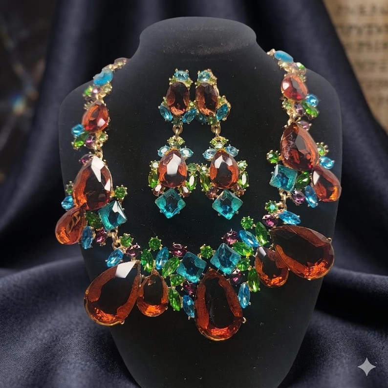 May include: A jewelry set consisting of a necklace and matching drop earrings. The pieces feature large, teardrop-shaped amber-colored crystals accented by smaller, faceted blue, green, and purple stones set in a gold-toned metal frame.
