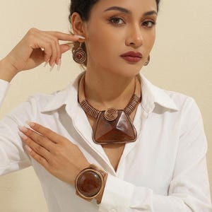 May include: A jewelry set consisting of a copper-toned collar necklace with a large trapezoidal brown stone pendant, matching spiral-patterned drop earrings, and a wide cuff bracelet featuring a large circular brown stone center piece.