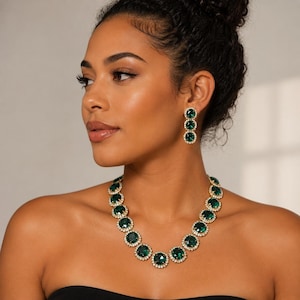 May include: A jewelry set consisting of a necklace and matching drop earrings. The necklace features a series of round, faceted emerald green stones set in gold-toned metal, each surrounded by a border of small, clear white crystals.