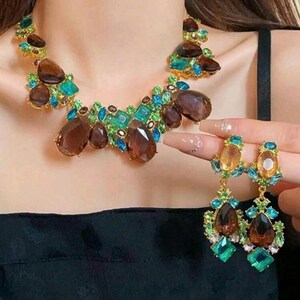 May include: A statement necklace and matching drop earrings featuring large, faceted brown teardrop stones, smaller bright turquoise and emerald green crystals, and clear accent gems set in a gold-toned metal base, creating a vibrant, multi-colored jewelry set.