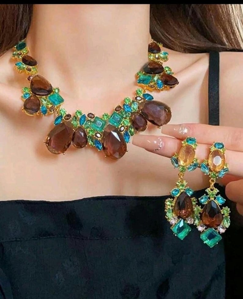 May include: A statement necklace and matching drop earrings featuring large, faceted brown teardrop stones, smaller bright turquoise and emerald green crystals, and clear accent gems set in a gold-toned metal base, creating a vibrant, multi-colored jewelry set.