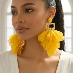 May include: Statement earrings featuring a small yellow beaded stud, a gold-tone rectangular frame, and a large, layered yellow fabric petal drop. The design incorporates a series of small, light-colored beads connecting the frame to the fabric petals.