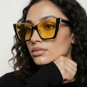 May include: Cat-eye sunglasses with a black frame and yellow lenses. The sunglasses have a gold accent on the top of the frame. The model is wearing a black top.