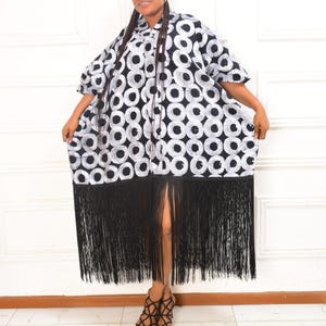 May include: A black and white patterned dress with a fringe hem. The dress features a repeating circle design and a collared neckline. The hemline is adorned with long black fringe. The person is wearing black strappy sandals.