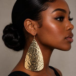May include: A large, teardrop-shaped earring with a metallic gold finish and a textured, hammered surface. The earring hangs from a simple hook wire, featuring a reflective, uneven surface that catches light across its wide, flat, pointed design.