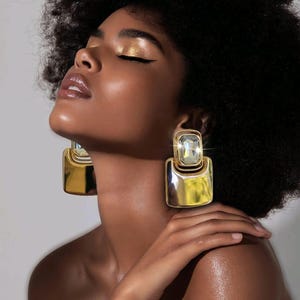 May include: Gold-toned square earrings with a clear, rectangular stone set in the center. The earrings have a polished, reflective surface. The image shows a close-up of the earrings, highlighting their design and the way they catch the light.