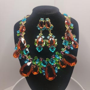 May include: A jewelry set consisting of a necklace and matching drop earrings. The pieces feature large, teardrop-shaped amber-colored crystals accented by smaller, vibrant turquoise, lime green, and purple faceted stones set in gold-tone metal.