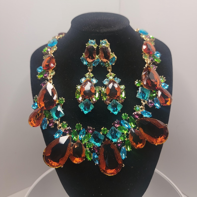 May include: A jewelry set consisting of a necklace and matching drop earrings. The pieces feature large, teardrop-shaped amber-colored crystals accented by smaller, vibrant turquoise, lime green, and purple faceted stones set in gold-tone metal.