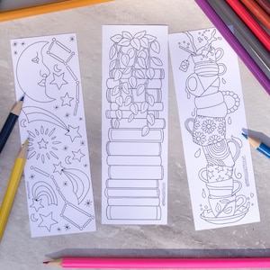 May include: Three white bookmarks with black line drawings of a moon and stars, stacked books with plants, and stacked teacups. Colored pencils in various colors surround the bookmarks.
