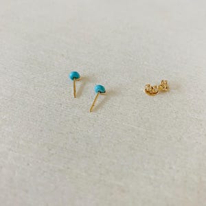 May include: Two gold stud earrings with small, round, turquoise stones. A gold earring back is also visible. The earrings are set against a light-colored background.