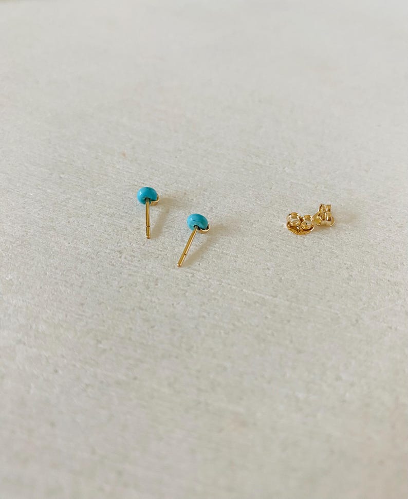 May include: Two gold stud earrings with small, round, turquoise stones. A gold earring back is also visible. The earrings are set against a light-colored background.