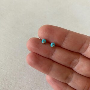 May include: A pair of small stud earrings. Each earring features a round, light blue bead with a gold-colored post. The earrings are displayed on a light background.
