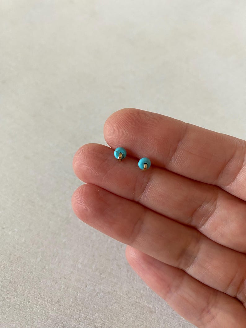 May include: A pair of small stud earrings. Each earring features a round, light blue bead with a gold-colored post. The earrings are displayed on a light background.