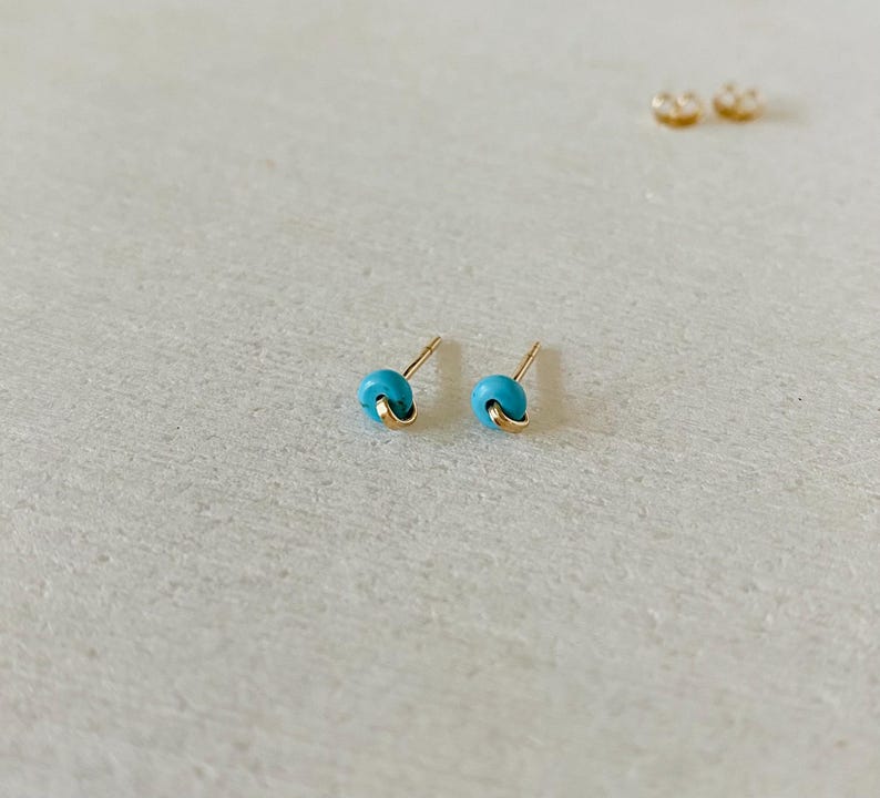 May include: A pair of small turquoise stud earrings with gold-colored accents. The earrings have a rounded shape and are set against a light background. Additional earring backs are visible in the background.