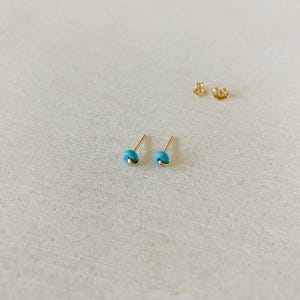 May include: A pair of small, gold-colored stud earrings with a turquoise bead. The earrings are displayed on a light-colored surface, with the earring backs visible in the background. The earrings have a simple, minimalist design.