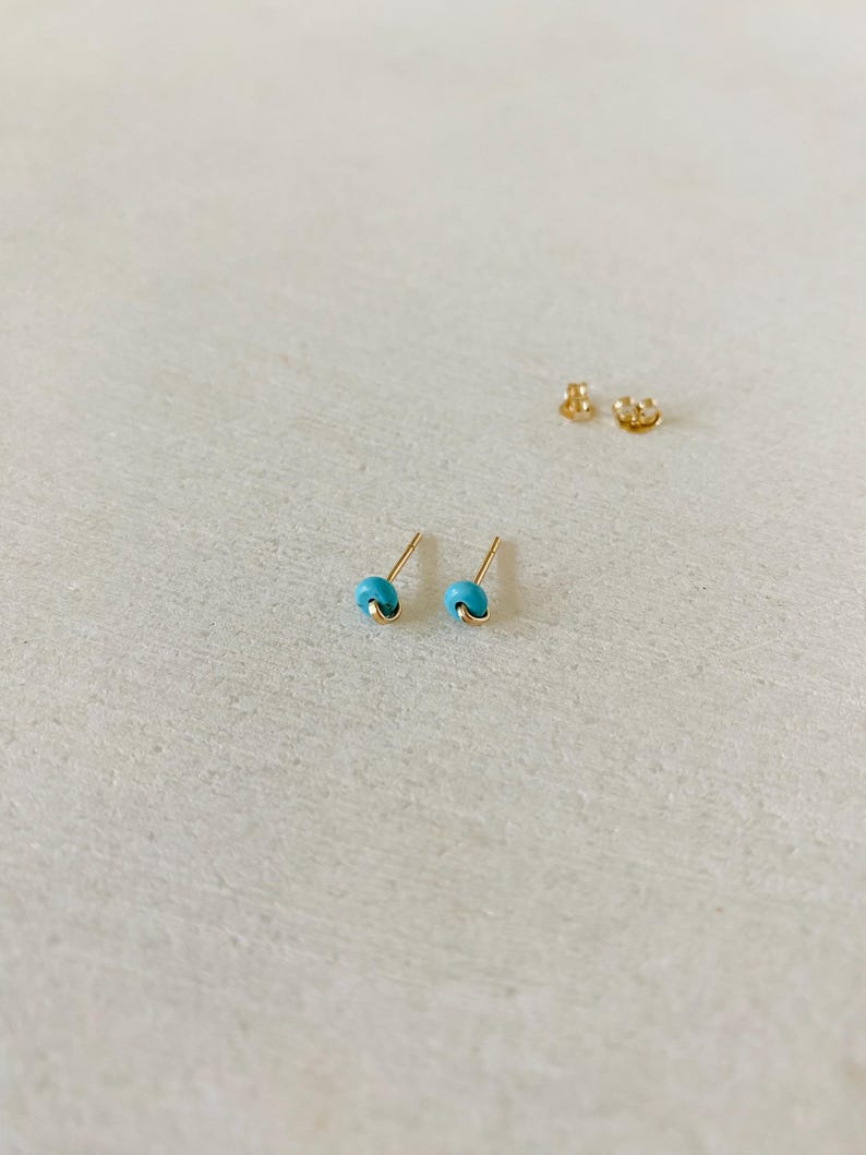 May include: A pair of small, gold-colored stud earrings with a turquoise bead. The earrings are displayed on a light-colored surface, with the earring backs visible in the background. The earrings have a simple, minimalist design.