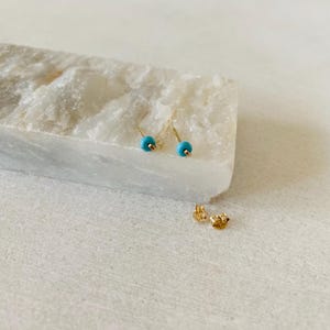 May include: A pair of gold stud earrings with small, round, turquoise-colored beads. The earrings are displayed on a white, textured surface, with two gold earring backs visible.