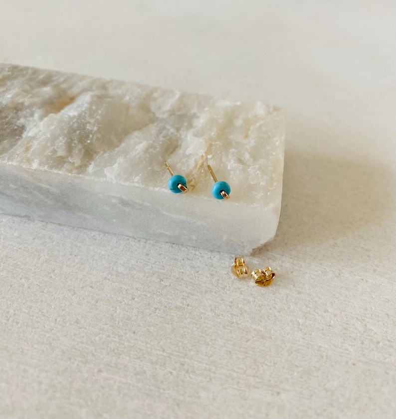 May include: A pair of gold stud earrings with small, round, turquoise-colored beads. The earrings are displayed on a white, textured surface, with two gold earring backs visible.