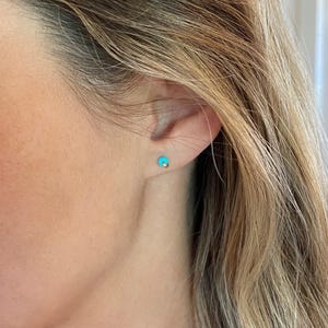 May include: A close-up of a person's ear with a small, turquoise stud earring. The earring has a gold post. The person has long, blonde hair.
