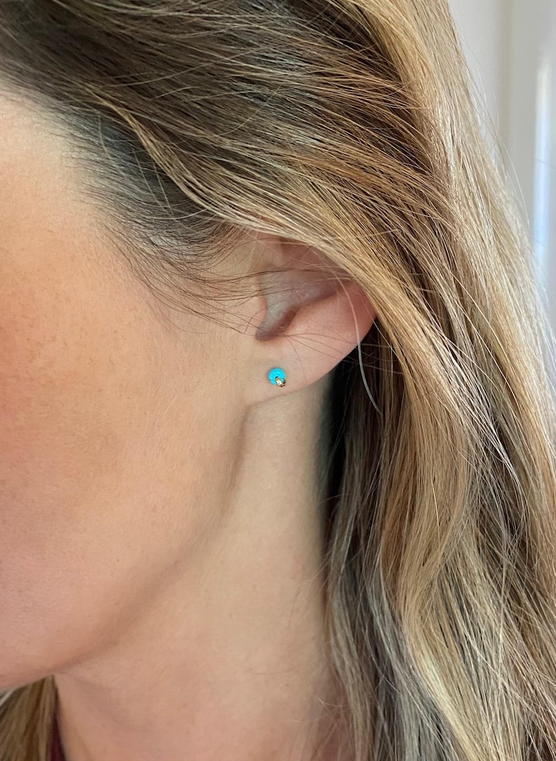 May include: A close-up of a person's ear with a small, turquoise stud earring. The earring has a gold post. The person has long, blonde hair.