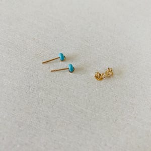 May include: Two gold-colored stud earrings with small, turquoise-colored stones. The earrings are displayed with a pair of earring backs on a light-colored surface. The earrings are small and delicate.
