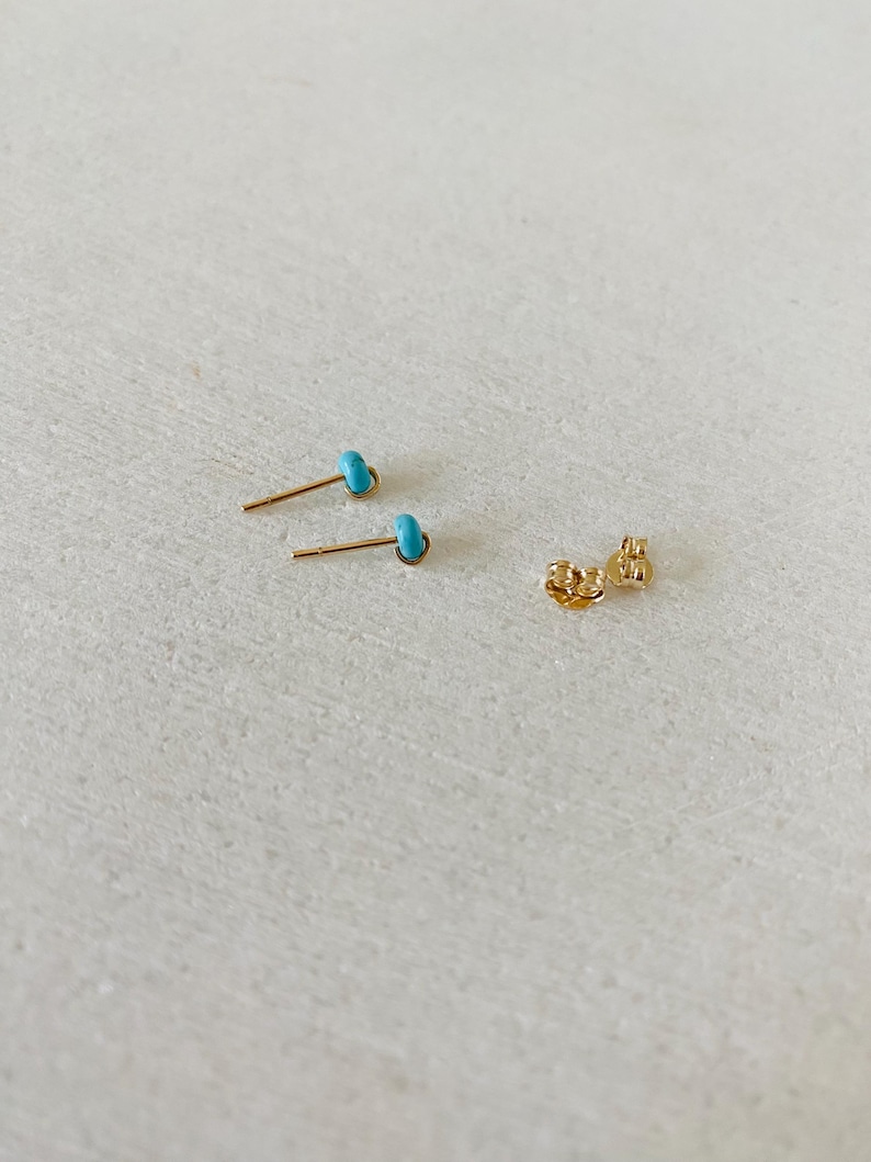 May include: Two gold-colored stud earrings with small, turquoise-colored stones. The earrings are displayed with a pair of earring backs on a light-colored surface. The earrings are small and delicate.