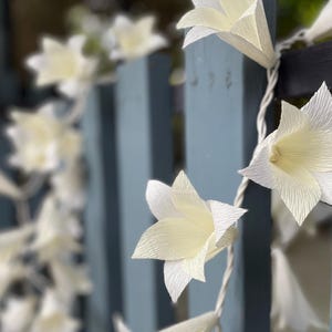 May include: A string of paper flower lights, featuring white and cream-colored lilies. The lights are strung on a white wire and are draped over a weathered, light blue wooden fence. The background is blurred.