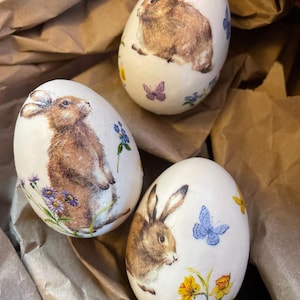 May include: Three white Easter eggs decorated with images of brown rabbits, butterflies, and flowers. The eggs are nestled in brown paper, creating a rustic and festive display. The eggs are a charming addition to Easter decor.