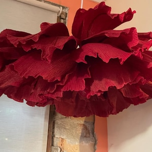 May include: A large, hanging decorative paper flower in a deep red hue. The flower has multiple layers of ruffled petals, creating a full, textured appearance. The paper is slightly crinkled, adding to the handmade aesthetic. The flower is suspended from a white string.