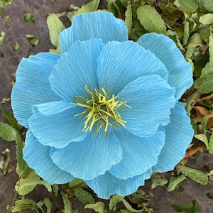 May include: A large, handmade, light blue paper flower with textured petals and a yellow center. The flower is surrounded by green leaves and rests on a dark, patterned surface, creating a striking contrast.