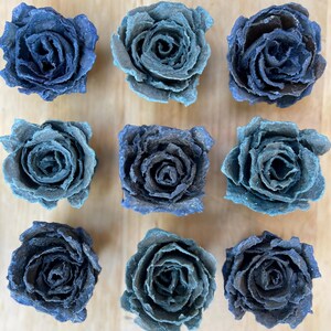 May include: Nine blue-toned paper roses arranged in a grid pattern. The roses vary in shades of blue, from light teal to deep indigo, with textured petals. The flowers are displayed on a light wooden surface.