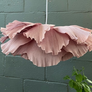 May include: A large, hanging paper flower in shades of pink. The flower has multiple layers of ruffled petals, creating a voluminous and textured appearance. The light fixture is suspended from a white cord.
