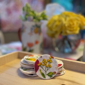 May include: A stack of round, white coasters with floral designs. The top coaster features a yellow flower with green leaves and red accents. The coasters are stacked on a light brown wooden tray, with blurred floral arrangements in the background.