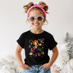 May include: A black short-sleeved t-shirt featuring a graphic print of the character Woody from Toy Story. The design includes a lasso, a sheriff star, horseshoes, a red boot, and the name Woody written in cursive script in a light color.