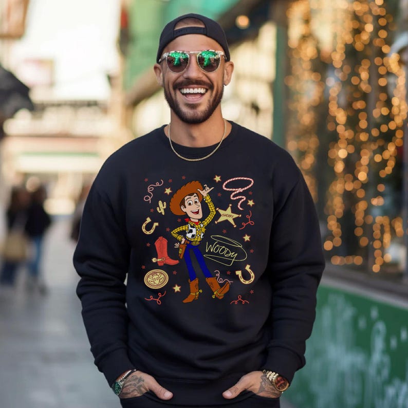 May include: A black long-sleeve sweatshirt featuring a graphic print of the character Woody from Toy Story. The design includes a central illustration of Woody surrounded by western-themed icons like a lasso, a sheriff star, a boot, and horseshoes.