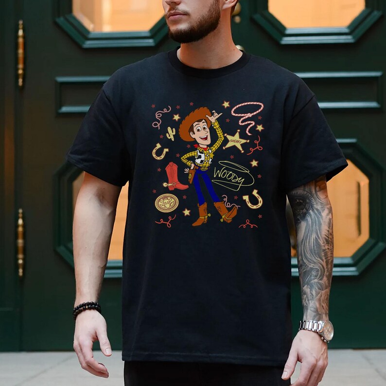 May include: A black short-sleeved t-shirt featuring a graphic of the character Woody from Toy Story. The design includes a central illustration of Woody surrounded by western-themed icons like a cowboy boot, a horseshoe, a star, and a lasso, with the name Woody written in a cursive script.