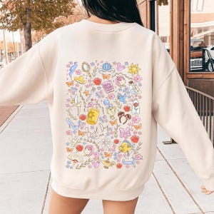 Puede incluir: Sudadera de color crema con un gráfico colorido de ilustraciones temáticas de cuentos de hadas, que incluyen un espejo mágico, una rosa y un zapato de cristal. Las palabras "Best Day Ever" están escritas en cursiva. El diseño está rodeado de flores y mariposas.
