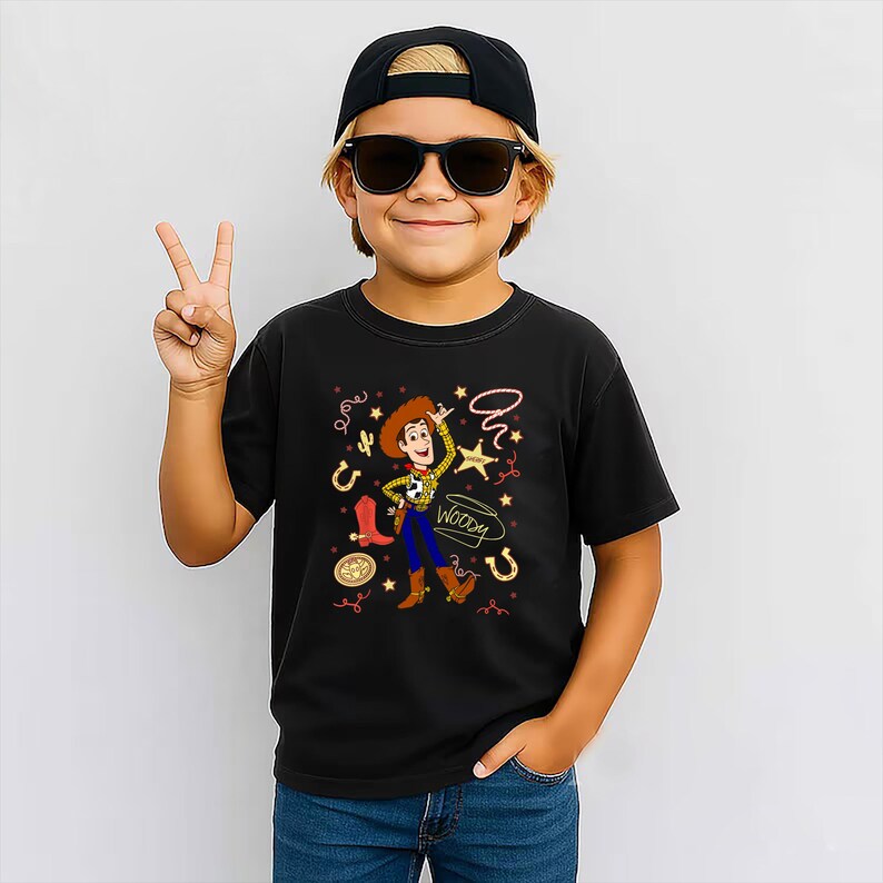 May include: A black short-sleeved t-shirt featuring a graphic print of the character Woody from Toy Story. The design includes a yellow plaid shirt, blue pants, brown boots, a cowboy hat, a sheriff star, a lasso, and the name Woody in script.