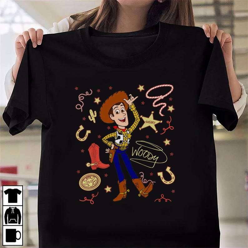 May include: A black short-sleeve t-shirt featuring a graphic of the character Woody from Toy Story. The design includes a sheriff badge, a lasso, a cowboy boot, a horseshoe, and the text Woody in a cursive script, surrounded by small stars.
