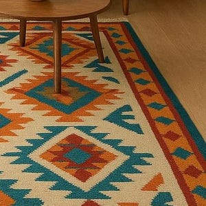 A multicolored area rug with a Southwestern or tribal geometric pattern is the centerpiece of a modern living room. The rug features bold diamond and zigzag shapes in shades of turquoise, orange, and blue on a light beige background, with a fringed border. It is placed on a light wooden floor in front of a neutral-colored sofa, a round wooden coffee table, and a large potted plant.