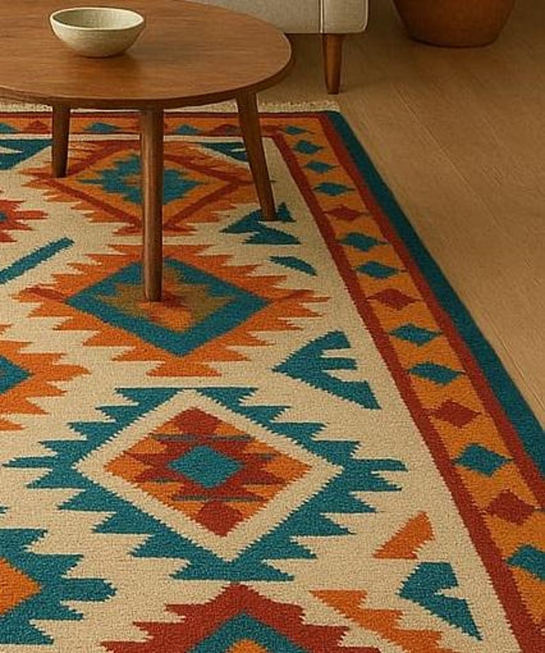 A multicolored area rug with a Southwestern or tribal geometric pattern is the centerpiece of a modern living room. The rug features bold diamond and zigzag shapes in shades of turquoise, orange, and blue on a light beige background, with a fringed border. It is placed on a light wooden floor in front of a neutral-colored sofa, a round wooden coffee table, and a large potted plant.