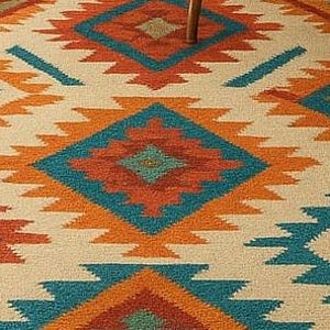 A multicolored area rug with a Southwestern or tribal geometric pattern is the centerpiece of a modern living room. The rug features bold diamond and zigzag shapes in shades of turquoise, orange, and blue on a light beige background, with a fringed border. It is placed on a light wooden floor in front of a neutral-colored sofa, a round wooden coffee table, and a large potted plant.