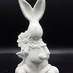 May include: White ceramic bunny figurine holding a flower. The bunny is wearing overalls and has a textured surface.