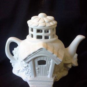 May include: White ceramic teapot shaped like a house with a flower-shaped roof and a small door. The teapot has a lattice window and a small porch.
