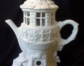 May include: White ceramic teapot shaped like a fairy house with a roof, windows, and a door. The teapot has a mushroom base and a small fence around the middle.