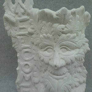 May include: White ceramic wall decor featuring a green man face with leaves and a welcome sign. The green man has a beard and mustache made of leaves. The welcome sign is made of a tree trunk with the word "welcome" carved into it.