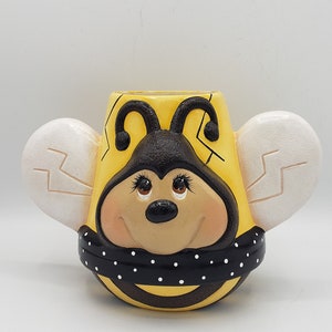 May include: A whimsical, hand-painted ceramic bee-shaped planter. The yellow body features a smiling bee face, black and white polka-dot scarf, and white wings. A charming decorative item for home or garden.